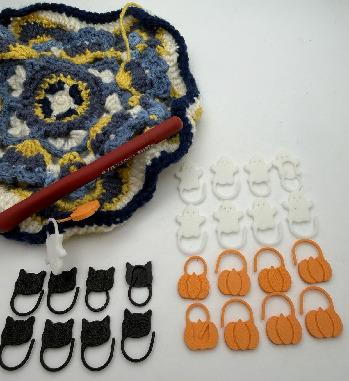 Halloween stitch markers in pumpkin, ghost and cat designs shown with a crochet project and hook