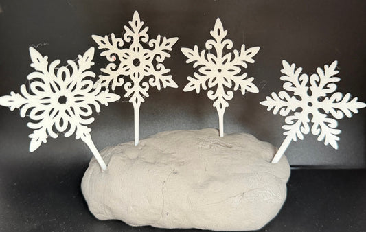 Four white filigree snowflake cake toppers standing in grey clay